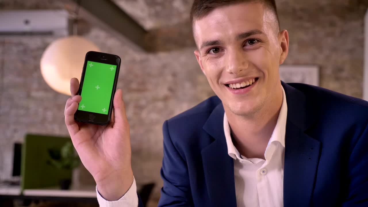 Young attractive businessman is watching smartphone in office, showing green screen, smiling, business concept, communication concept