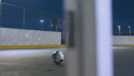 player kicks soccer ball toward goal post during intense urban night practice, ball rolls out after hitting goal, surrounded by illuminated city buildings