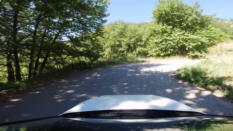 driver pov from a white car turning right on mountain road with reflective windshield and bright sunlight shining through green trees in countryside of southern europe