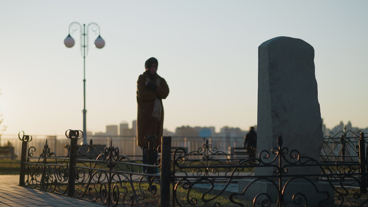 코트와 스카프를 입은 여자가 해가 지는 동안 공원에서 돌로 된 기념비 앞에 서 있습니다. 이 장면은 는 태양의 따뜻한 빛으로 긴 그림자를 던집니다.