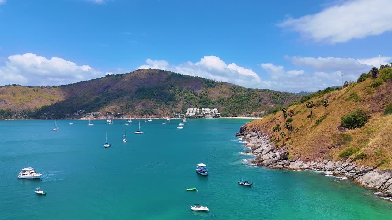 푸켓의 거친 해안선을 따라 푸르키즈색 물에 있는 보트를 찍은 공중 영상, 은 하늘 아래, 평온한 자연의 아름다움을 보여줍니다.