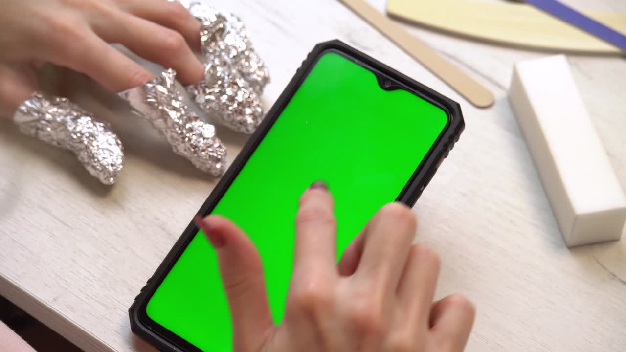 매니큐어 중 한 소녀는 녹색 화면을 가진 스마트 폰을 사용합니다. 자체 매니큐르 서비스. 매니큐리스트는 분홍색 젤 폴라시로 손을 칠합니다. 매니쿠어 된 빨간 손. 손 폴라시 응용 프로그램.