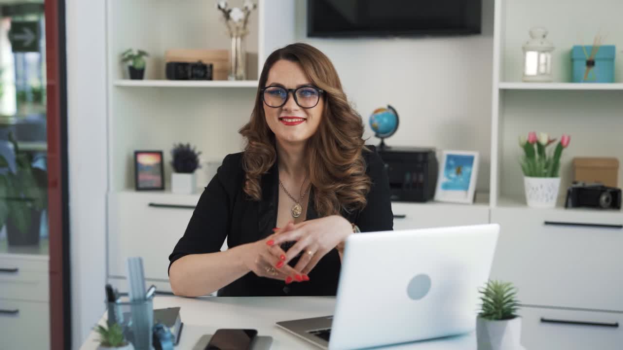 The portrait of a middle-aged woman sitting at a desk in front of a laptop and smiling, dressed in business style and wearing glasses for vision