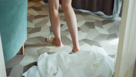 woman wearing white robe and soft white slippers walking slowly toward curtain across patterned geometric floor, robe swaying gently beside teal chair in softly lit serene interior