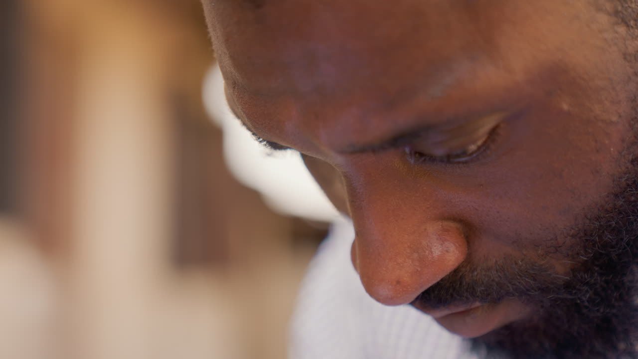 Homem negro em close-up extremo lendo, foco editorial intenso com testa franzida e barba, luz quente cinematográfica, detalhe macro na textura da pele e nos olhos, clima de determinação durante a revisão de texto.