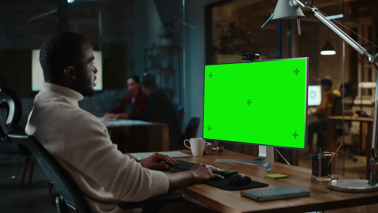 el guapo gerente de proyecto afroamericano está haciendo una llamada de video en una computadora de escritorio con pantalla verde en una oficina creativa ocupada.