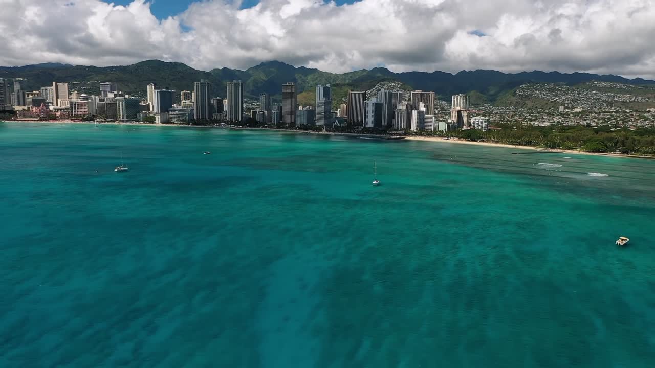 Aerial drone view showing North Shore of Oahu with turquoise ocean water, sandy beach, green mountains, and distant city skyline under partly cloudy sky in Hawaii