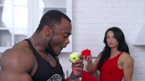 fit people eating healthy food. big strong african man enjoying apple. woman eating red pepper at the backdrop in blur. white room background.