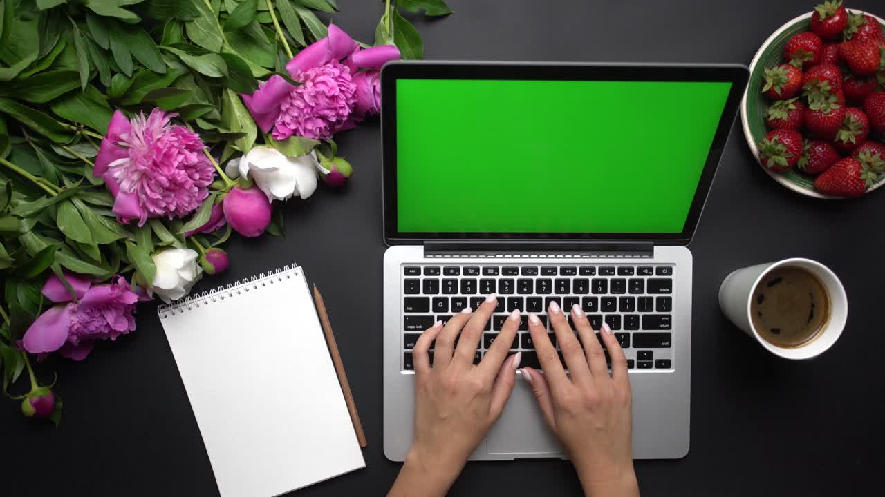 Female workspace. Woman tapping on the laptop computer keyboard with green screen. Bouquet of peony flowers, notebook and coffe on black background. Chroma key. Top view