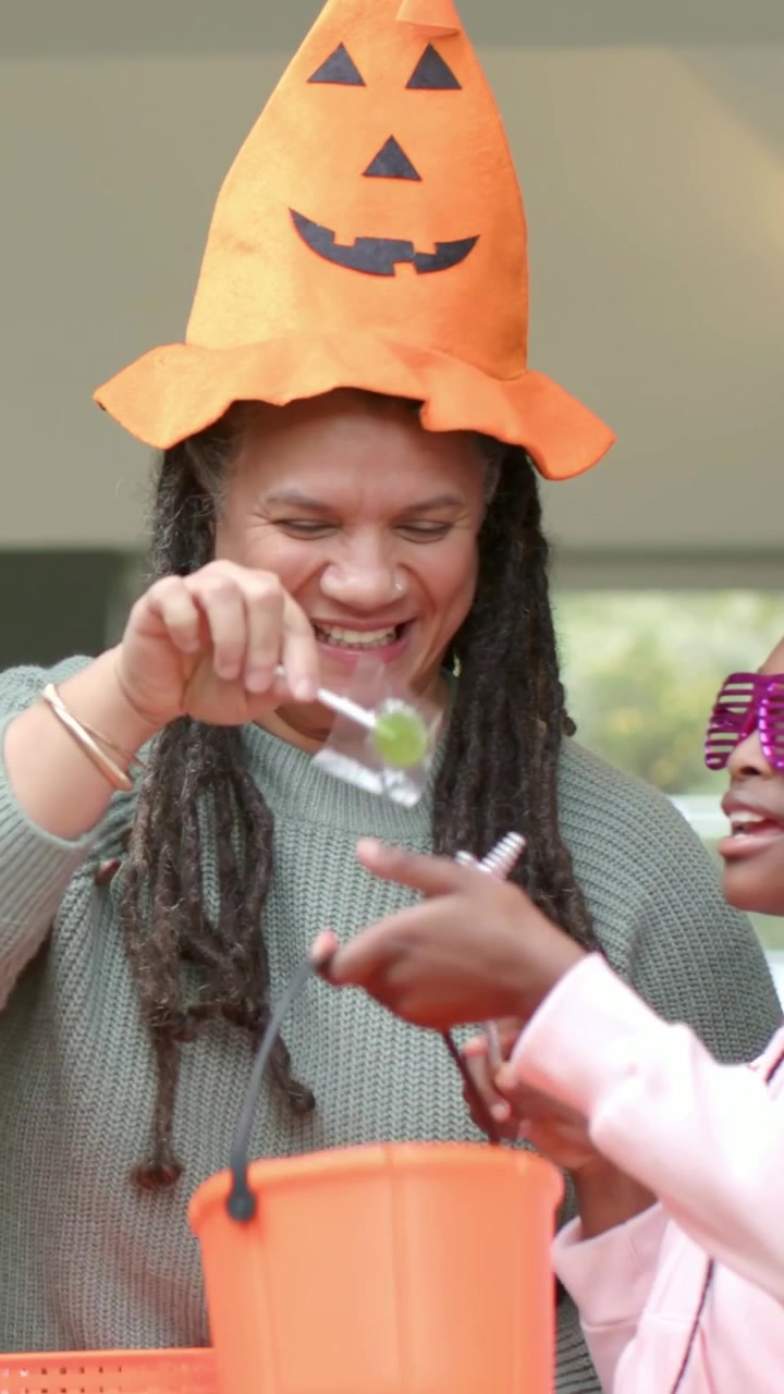 Vertical video: Child reaching from right causing pumpkin hat woman handing candy at porch, bucket