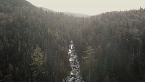 Arroyo-Que-Fluye-Suavemente-A-Través-Del-Camino-De-Roca-Helada-Desde-La-Exuberante-Montaña-Del-Bosque-En-Vallee-Bras-du-nord,-Quebec,-Canadá---Toma-Aérea-De-Drones