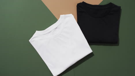 video of flat lay of folded white and black t shirts with copy space on green and brown background