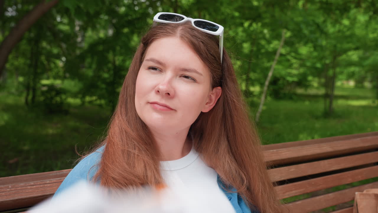 mujer elegante sentada en un banco del parque abanicándose la cara en un día soleado, con los ojos cerrados, el pelo ondeando, aliviada por el clima cálido, con un fondo verde al aire libre, y con una camisa blanca informal y una sobrecamisa azul