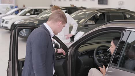 young car salesman showing to young couple new automobile at dealership salon.