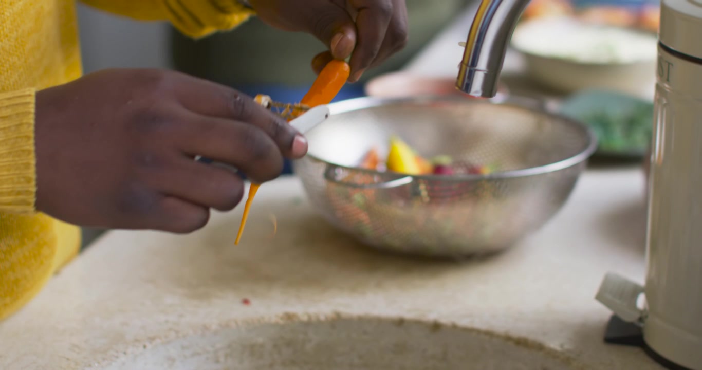 Casal na cozinha, homem descascando cenoura com descascador branco, preparando o jantar enquanto a mulher segura uma caneca.