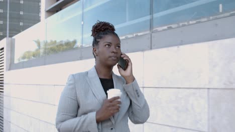 dolly shot of a confident african-american businesswoman walking outside, holding coffee and talking on mobile phone