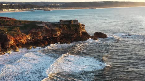 El-Faro-De-Nazare-Portugal-En-Praia-Do-Norte-Durante-La-Puesta-De-Sol-De-La-Hora-Dorada,-Muñeca-Aérea-Desde-Un-Lado