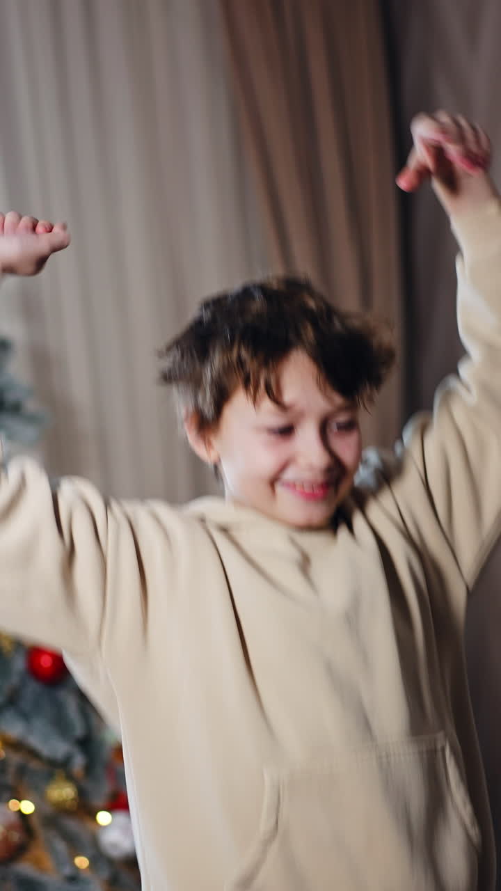 Happy rejoicing Caucasian kid in beige hoodie in the room. Dark-haired teenager jumps happily at the backdrop of Christmas tree. Vertical video