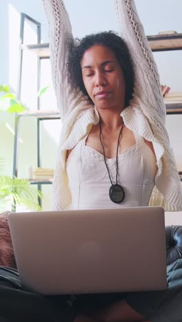 young african american woman stretches hands up sits on sofa with laptop on lap