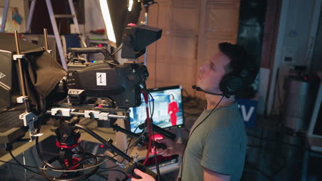 cameraman with headset operates professional broadcast camera in television studio while recording female presenter in red dress standing on platform in front of blue screen during video production session