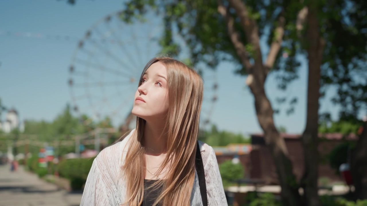 Close up of young woman with long brown hair wearing light colored cardigan and black top standing outdoors in park like setting. Background features trees clear blue sky distant amusement