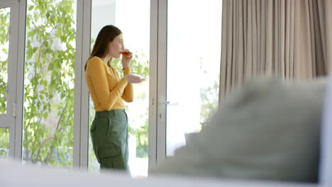 thoughtful caucasian woman drinking tea and looking through window at home, copy space, slow motion