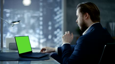 Young and Perspective Businessman Sits at His Table and Types on a Laptop with Green Mock-up Screen. Her Office Looks Modern with Dark Overtones, Big City Behind the Window.