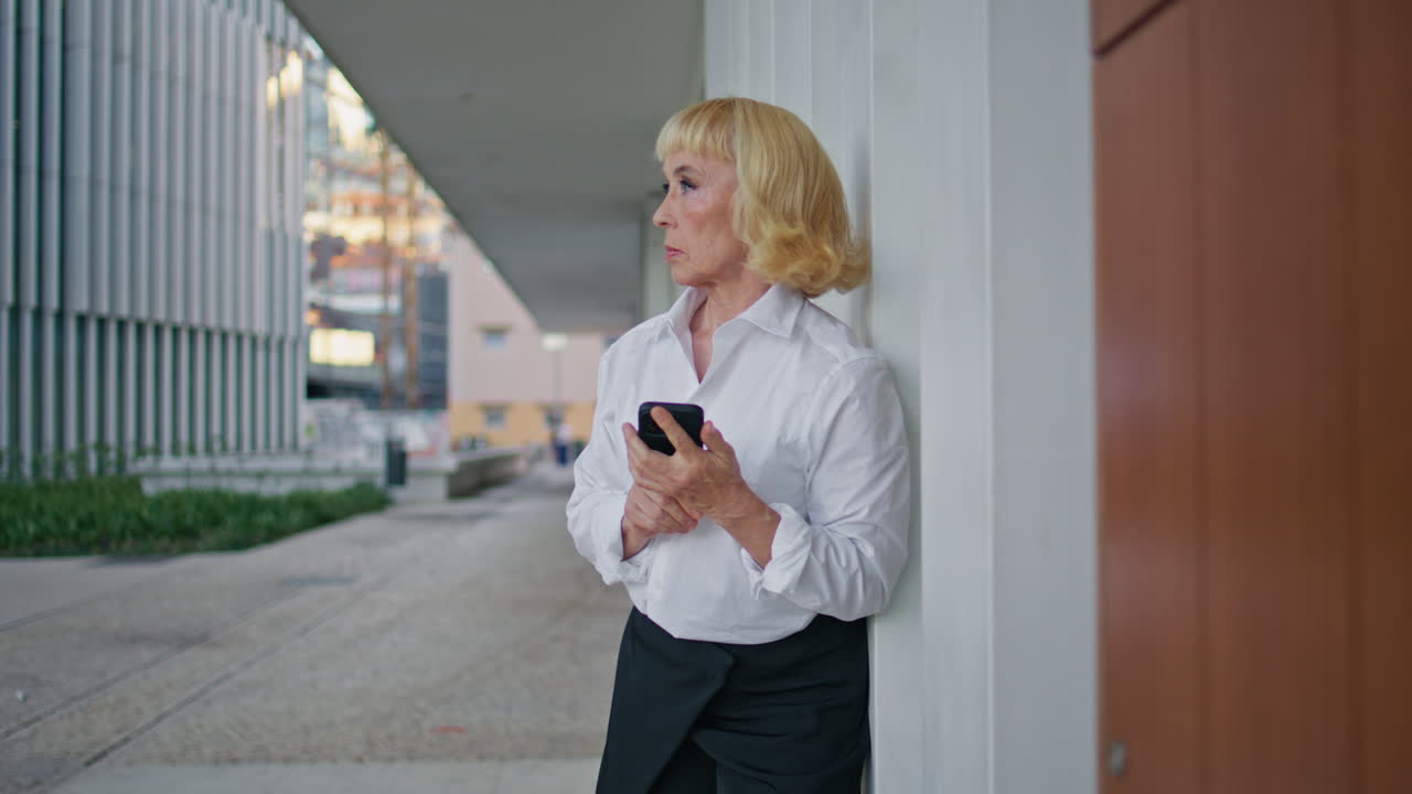 Aged businesswoman waiting meeting holding smartphone on street. Elderly woman