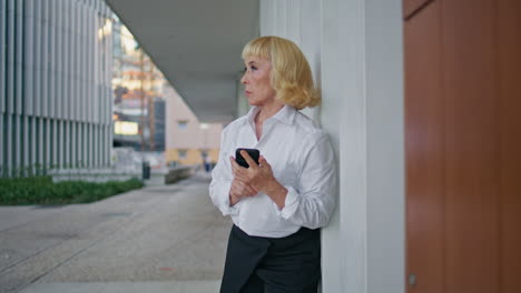 aged businesswoman waiting meeting holding smartphone on street. elderly woman