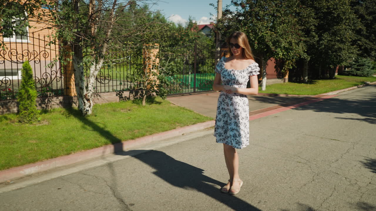 Thoughtful woman in floral dress and sunglasses walks with hands together under bright sun, passing residential property with iron fence, trees casting shadows on quiet sidewalk and street