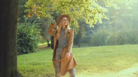 cheerful blonde young woman wearing hat and coat with backpack running and whirling in the park in autumn