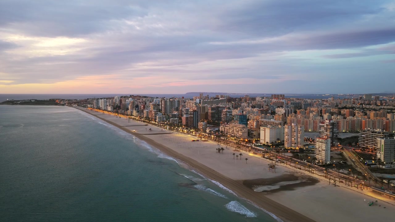 Città illuminata di Alicante e la sua lunga spiaggia sabbiosa durante il crepuscolo colorato dell'ora blu in Spagna. Drone aereo in ascesa, spazio per il testo.