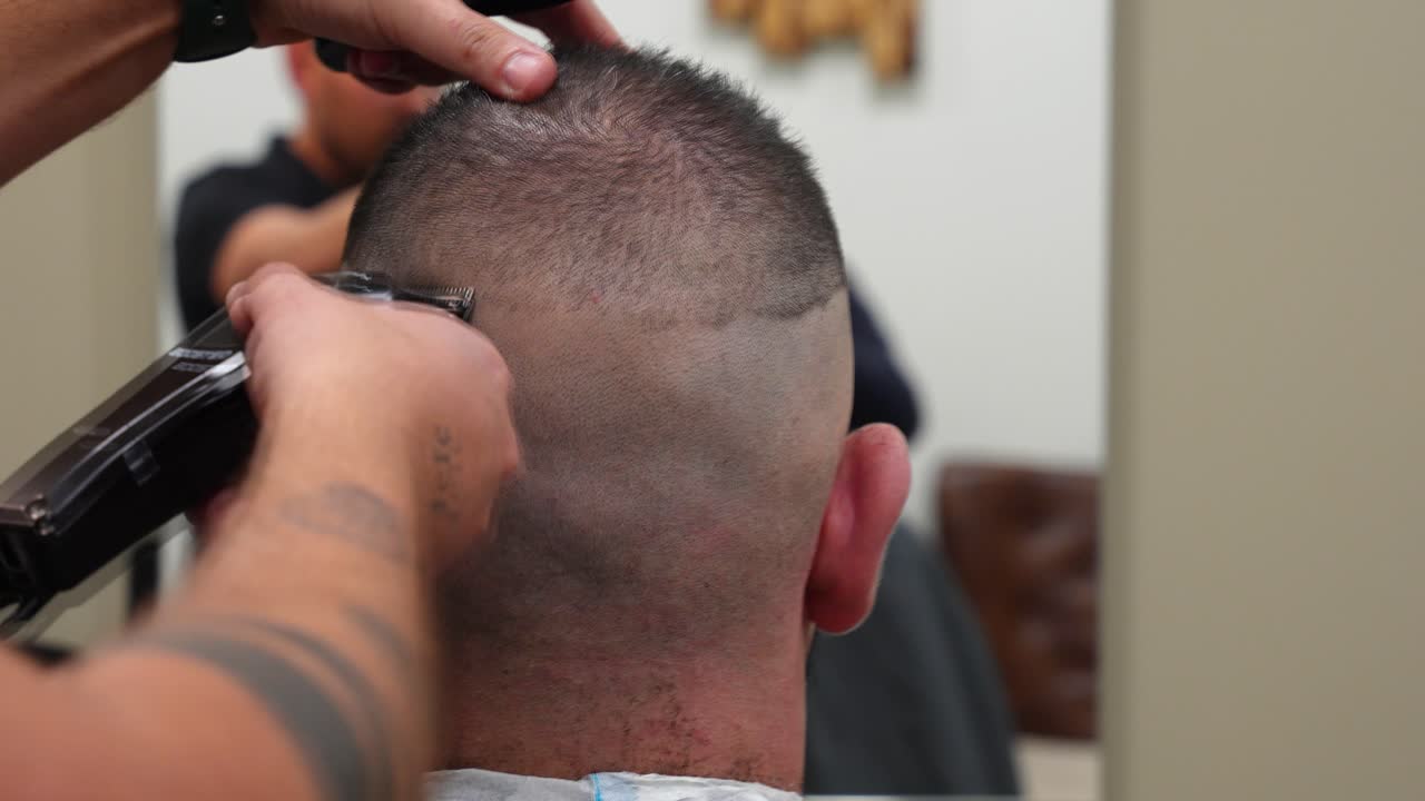 Slow motion rear view of a professional hairdresser shaving a man's head with a trimmer in barbershop