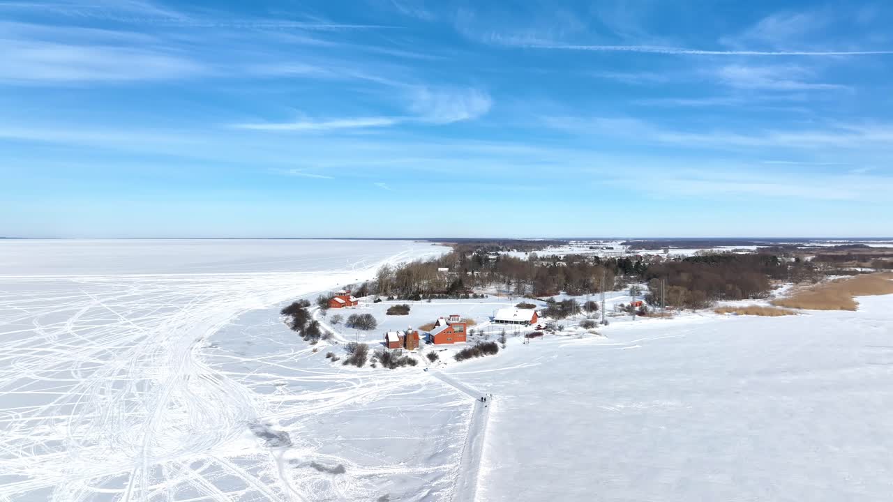 Wide-angle drone shot capturing the entire snowy peninsula of Ventė Horn, featuring red buildings and a lighthouse surrounded by a vast frozen lagoon