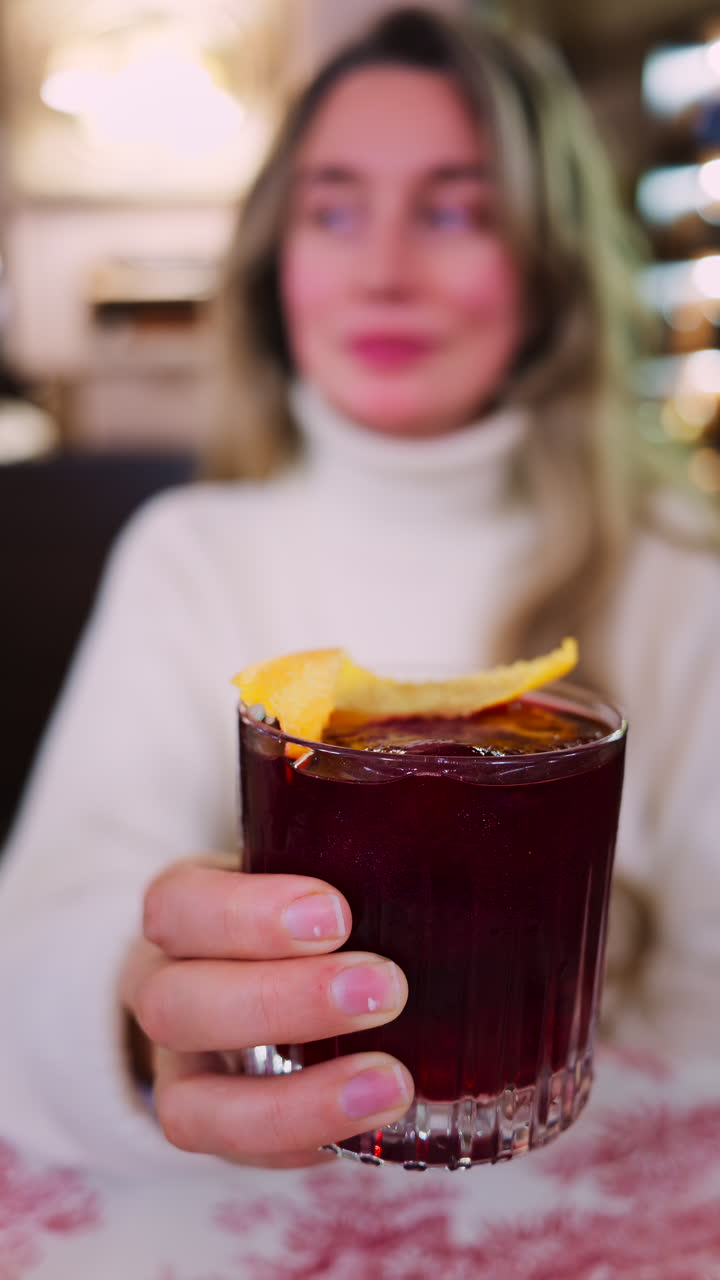 Close up of a woman holding a negroni cocktail on a red and white tablecloth at a restaurant. Vertical