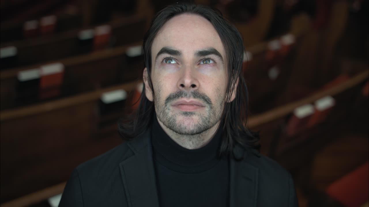 A Christian man, priest, preacher, pastor smiles in a black turtleneck and blazer stands in a church sanctuary, framed by wooden pews and soft light, creating a cinematic and contemplative mood