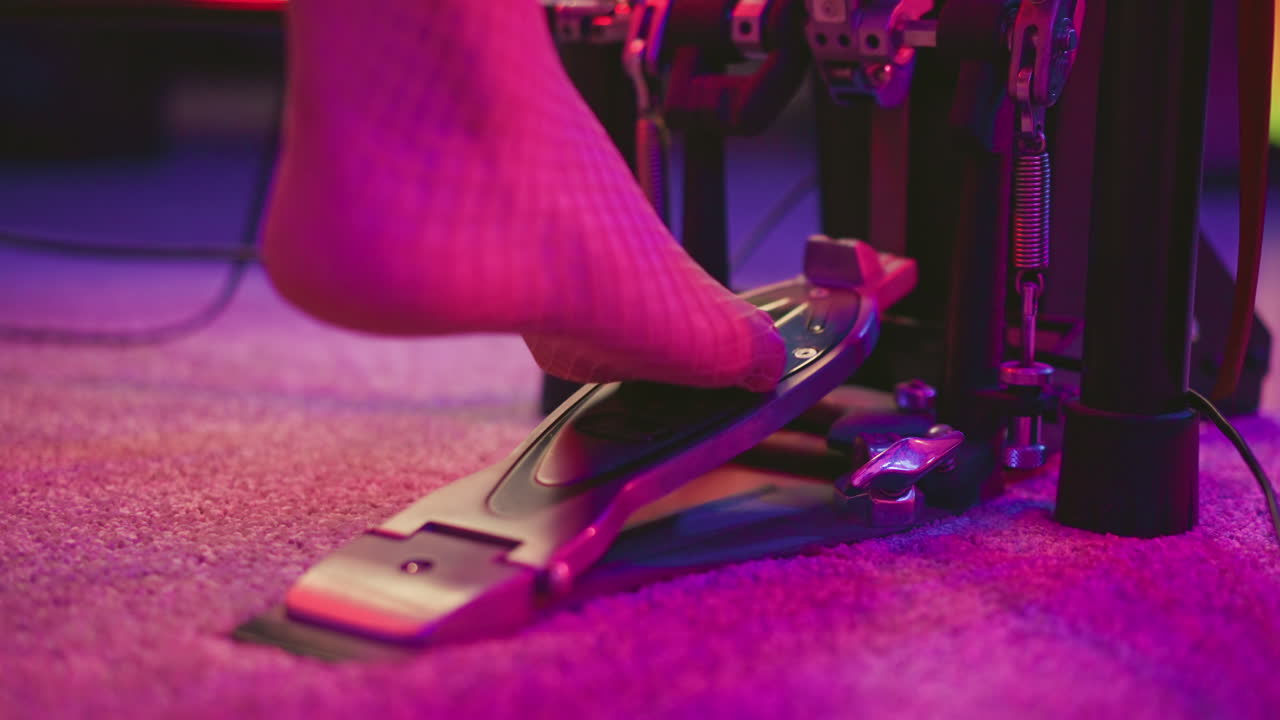 Close up of bare foot in fishnet pantyhose pressing kick drum pedal during music practice on carpeted floor under neon pink and purple lighting in studio setting with mechanical pedal details in view