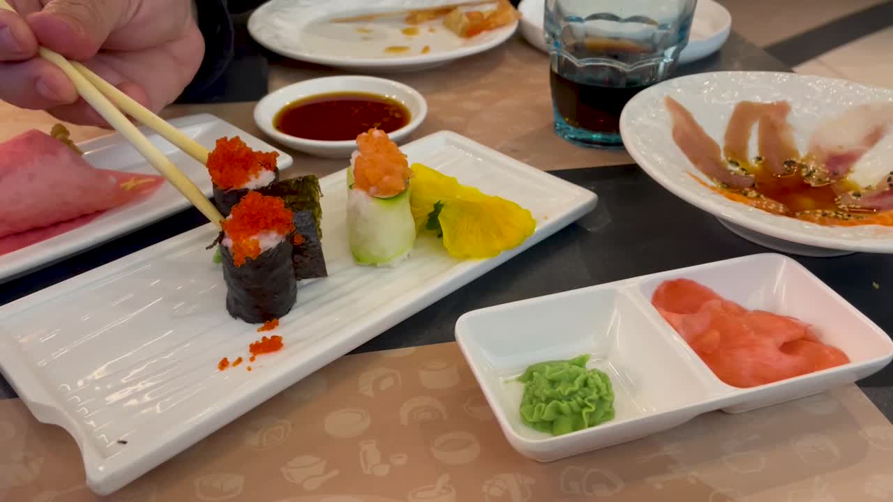 vemos una mesa con platos asiáticos, una con wasavi y jengibre, una con carpaccio de pescado, un temaki y sushi, una mano que aparece con palillos tomando sushi y sumergiéndolo en salsa de soja