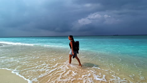 woman with backpack walks barefoot along turquoise shoreline as storm clouds gather over gili trawangan, indonesia