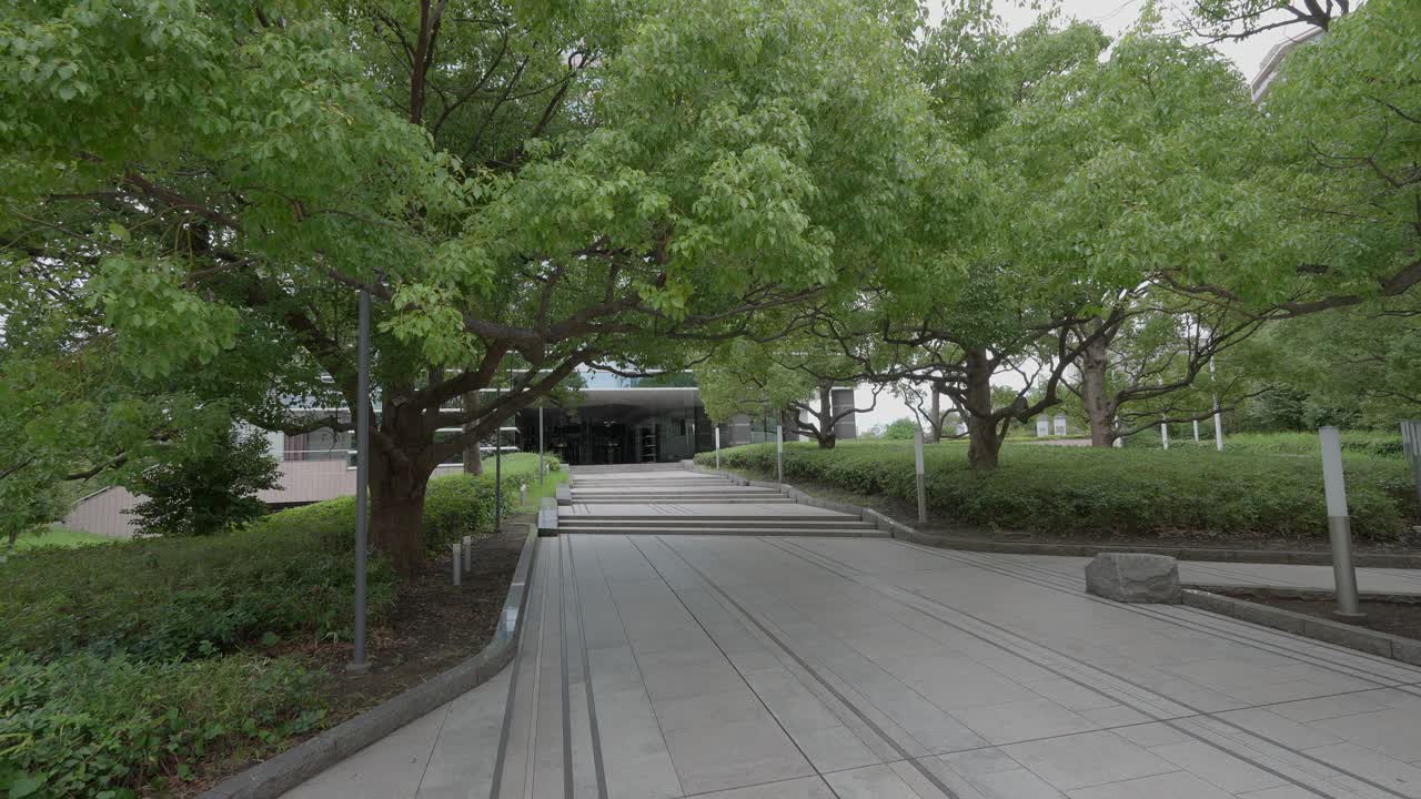 A peaceful shot of a paved walkway leading toward a modern building, surrounded by green trees and foliage