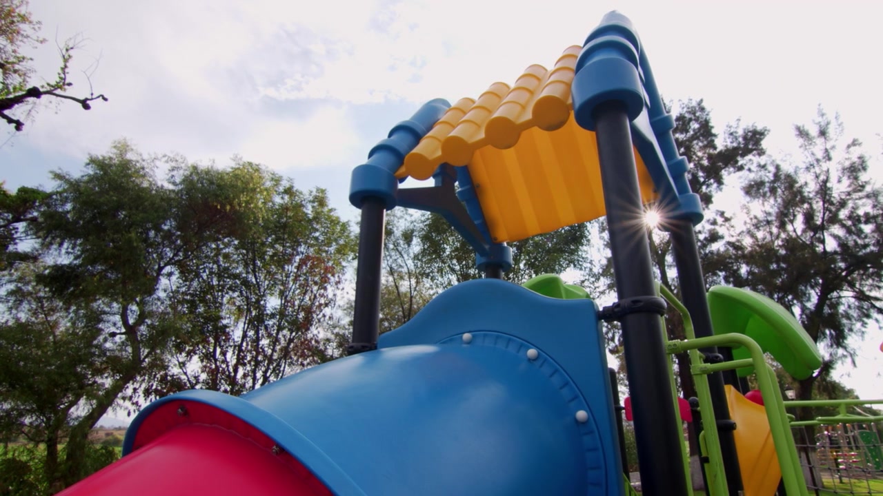 Primer plano de un tobogán de tubo colorido en un parque infantil soleado, con equipos de juego azules y amarillos, árboles y espacio de recreación al aire libre