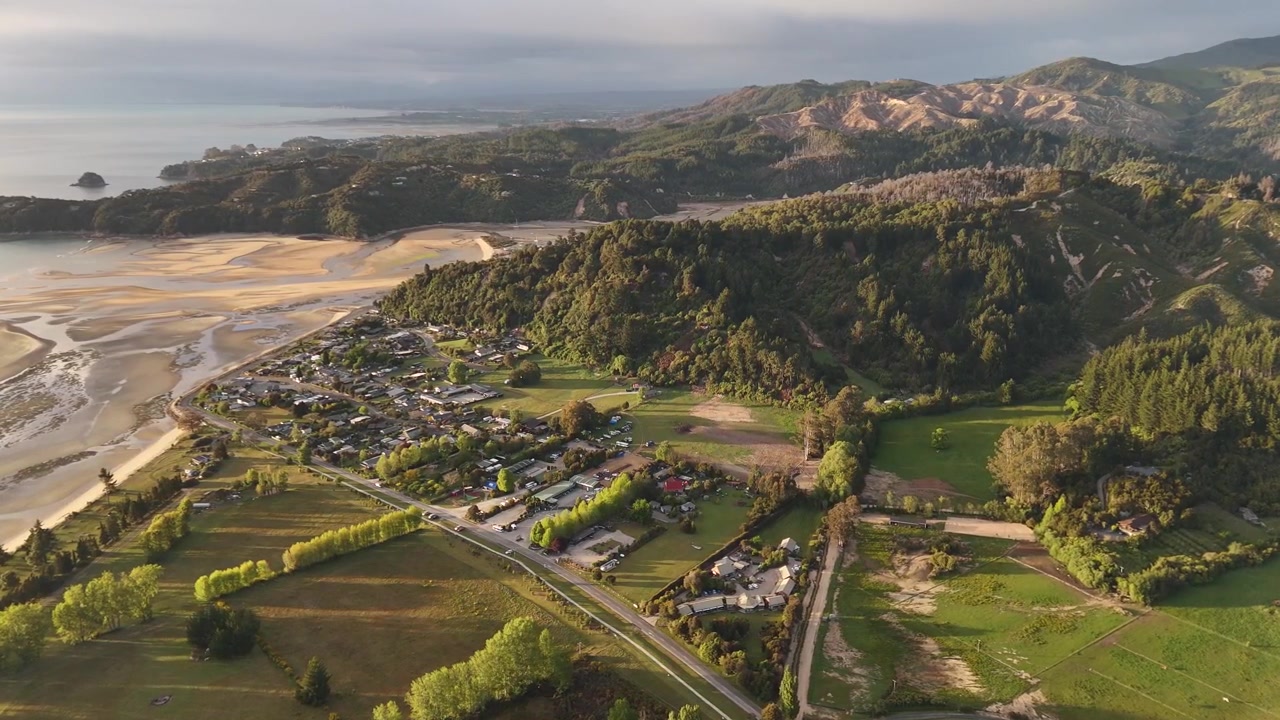 Yeni Zelanda'nın Güney Adası'ndaki Abel Tasman Milli Parkı'nın kenarında, yemyeşil tepeler ve altın rengi sabah ışığıyla gün doğumunda Marahau köyü ve kıyı haliçi. Havadan görünüm.
