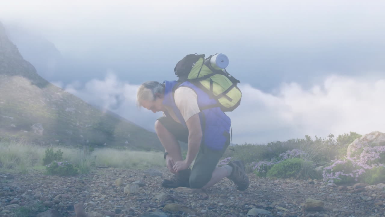 Mature male hiker kneeling tying boot laces on mountain trail, showing travel backpack overlay