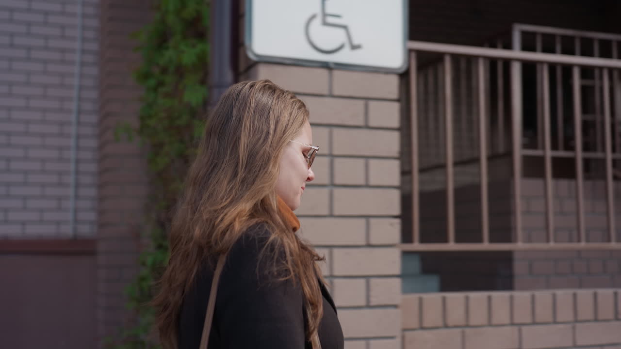 Vista lateral de un estudiante en el campus con cabello castaño claro y gafas de sol caminando junto a una pared de ladrillo y una barandilla en una tranquila mañana de ciudad con una suave brisa que levanta mechones y una correa de bolso visible