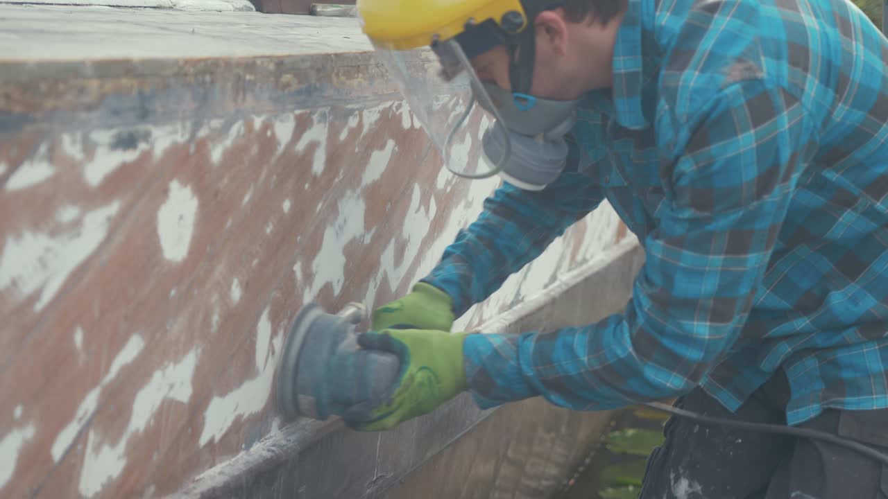 un carpintero que pinta vieja pintura en el casco de un barco de madera usando un licuador