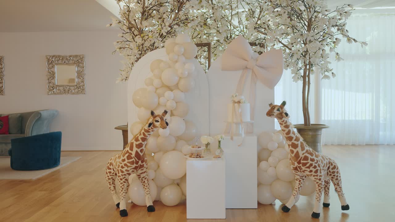 Elegante montaje para un baby shower con arreglos de globos blancos, iluminación suave, detalles florales y peluches de jirafas en un espacio interior luminoso