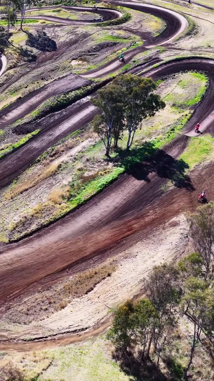 Drone segue piloto de motocross navegando por uma pista sinuosa, dia ensolarado, paisagem natural, movimento suave.