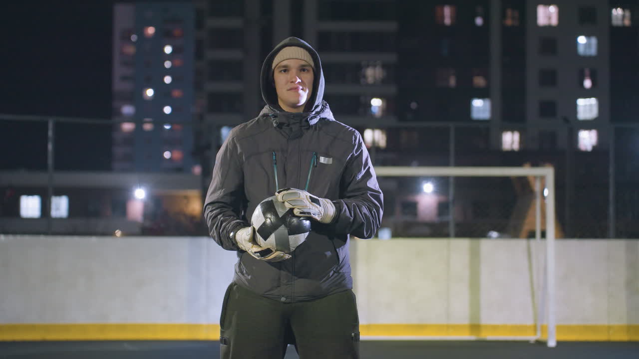 Man in hoodie and gloves holding soccer ball thoughtfully under night lights on urban sports field, high-rise buildings and goalpost in background