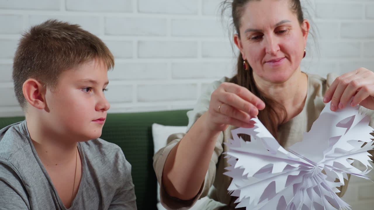 mother proudly unfolding intricate paper craft while son watches closely with amazed expression sitting together on green sofa in cozy room decorated with christmas tree and white brick wall