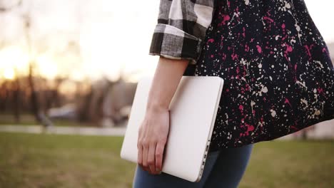 View-from-the-side-of-the-girl-in-jeans-walking-through-the-park-with-a-colored-bag-and-a-laptop-in-hand.-Close-up